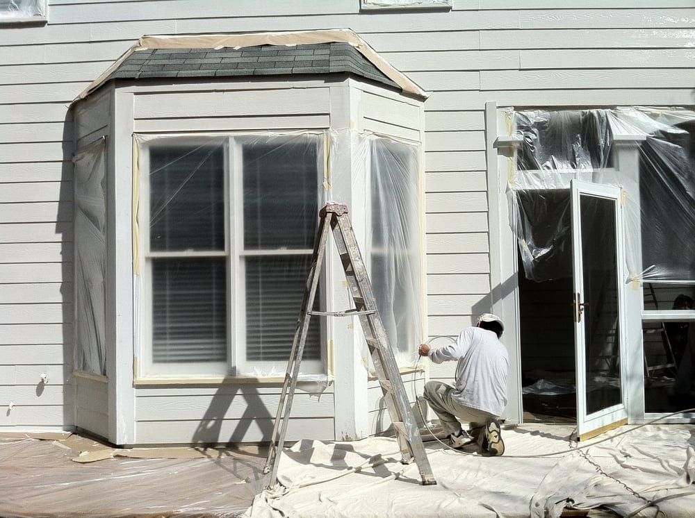 Exterior with a bay window, a ladder, and plastic over the door while a painter preps the siding