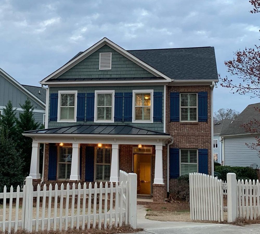The same blue-gray brick house at dusk with porch lights on and a white picket fence