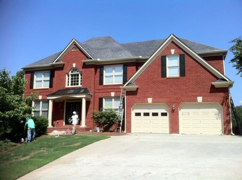 Red brick two-story house with black shutters and two garage doors, with painters by the front door