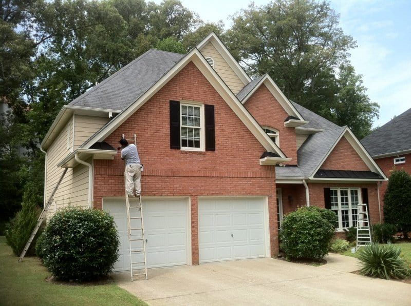 Red brick house with cream siding, and a painter on a ladder at the gable above the garage