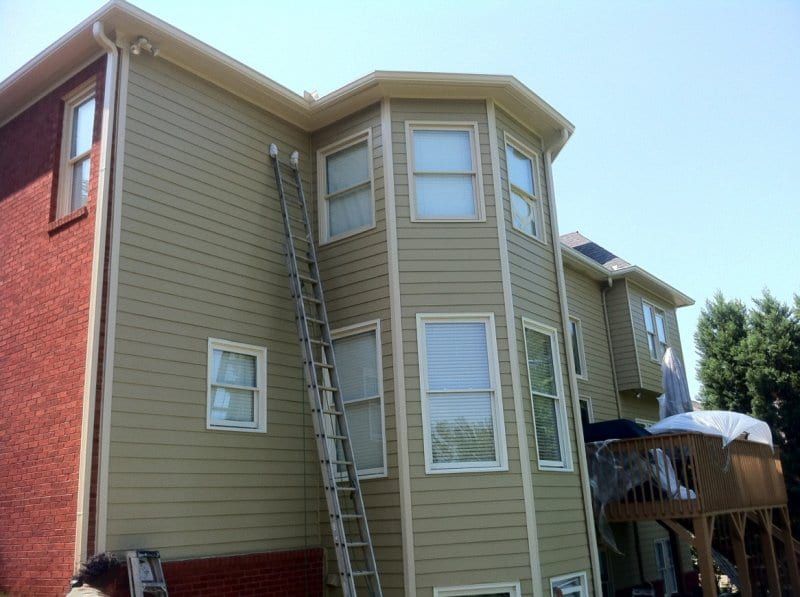Tall house with tan siding and a brick base, with a ladder at the bay window