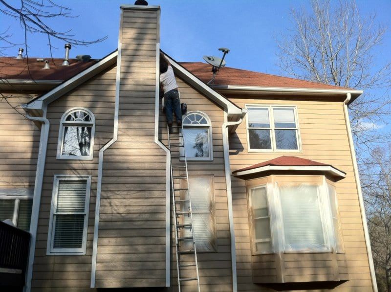 Painter on a ladder beside the chimney of a tan sided house