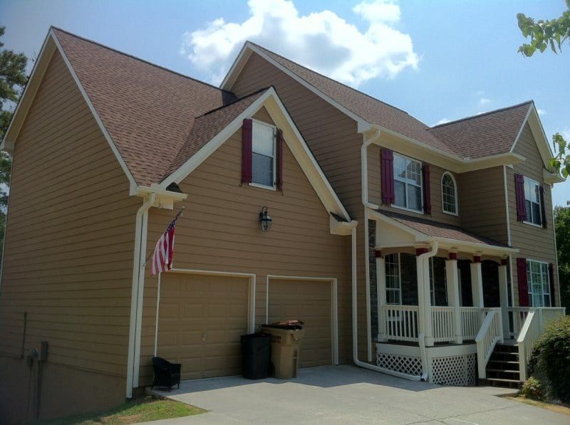 Tan sided house with a front porch, red shutters, and an American flag