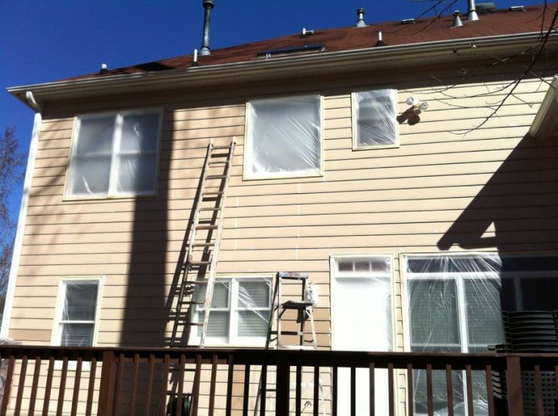 Back of a tan sided house with ladders against the wall above a deck