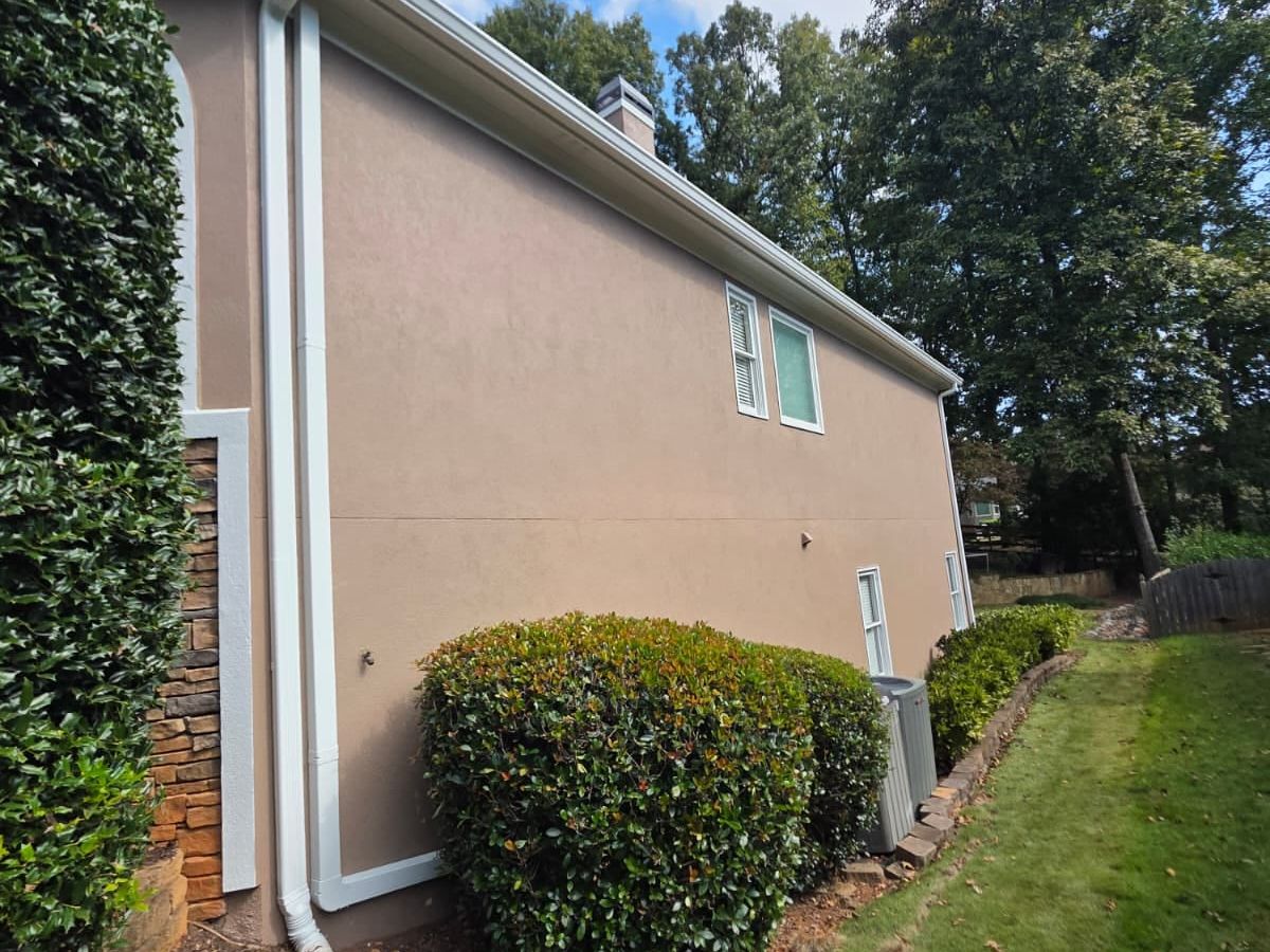 Side of a tan stucco house with white downspouts beside a green hedge